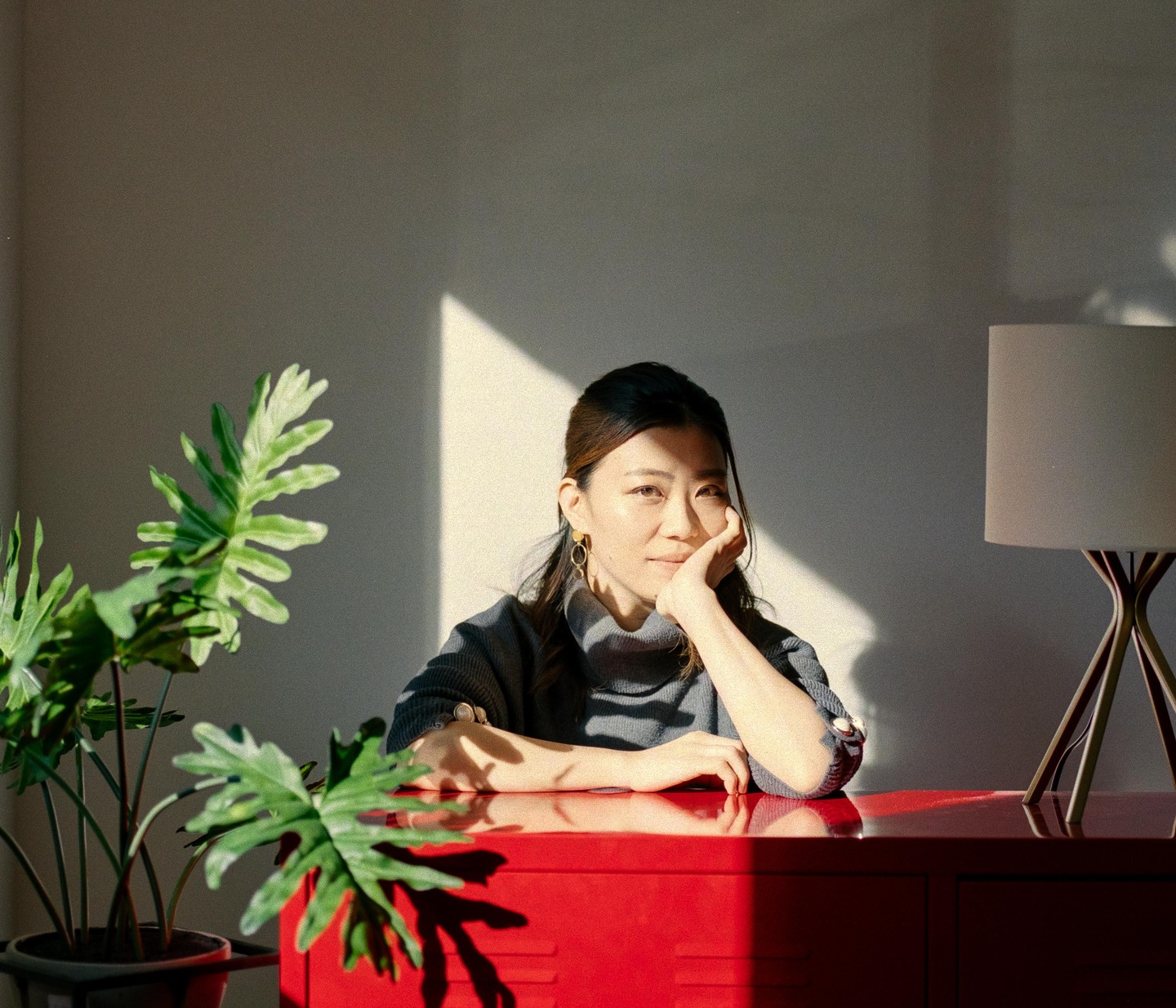 A woman rests her chin on her hand while sitting at a red desk with a plant and a lamp.