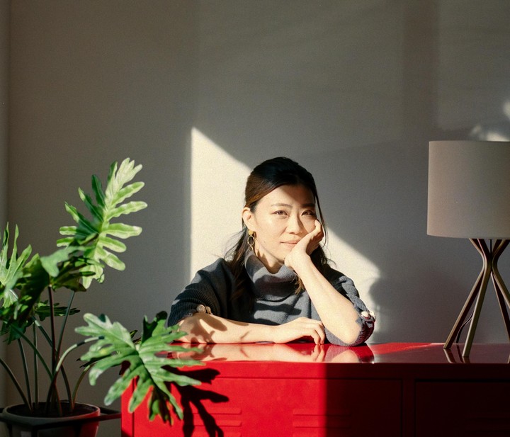 A woman rests her chin on her hand while sitting at a red desk with a plant and a lamp.