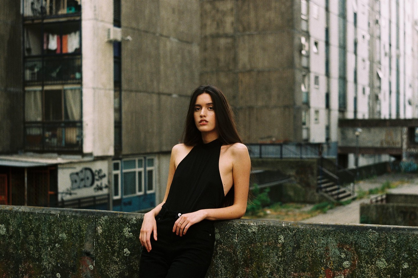 A woman with long dark hair leans against a mossy concrete wall in front of a large concrete building.