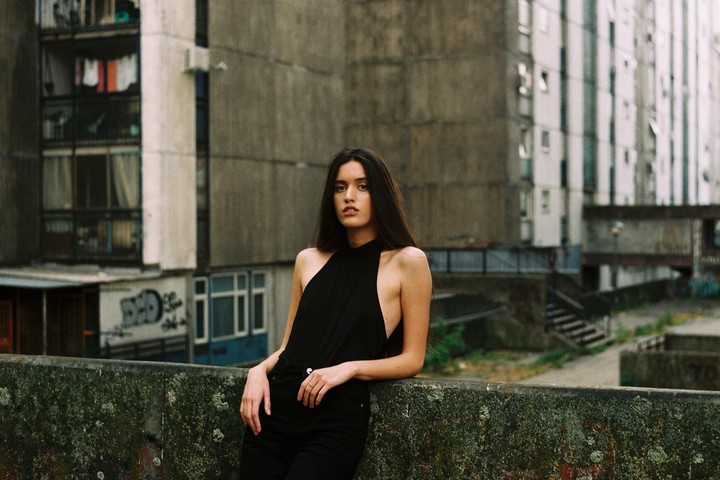 A woman with long dark hair leans against a mossy concrete wall in front of a large concrete building.
