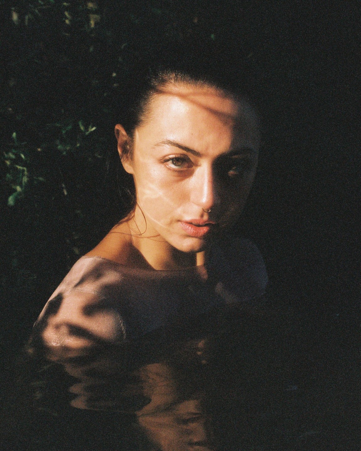 A woman with wet hair and shoulders is partially submerged in water, with dappled sunlight on her face and shoulder.