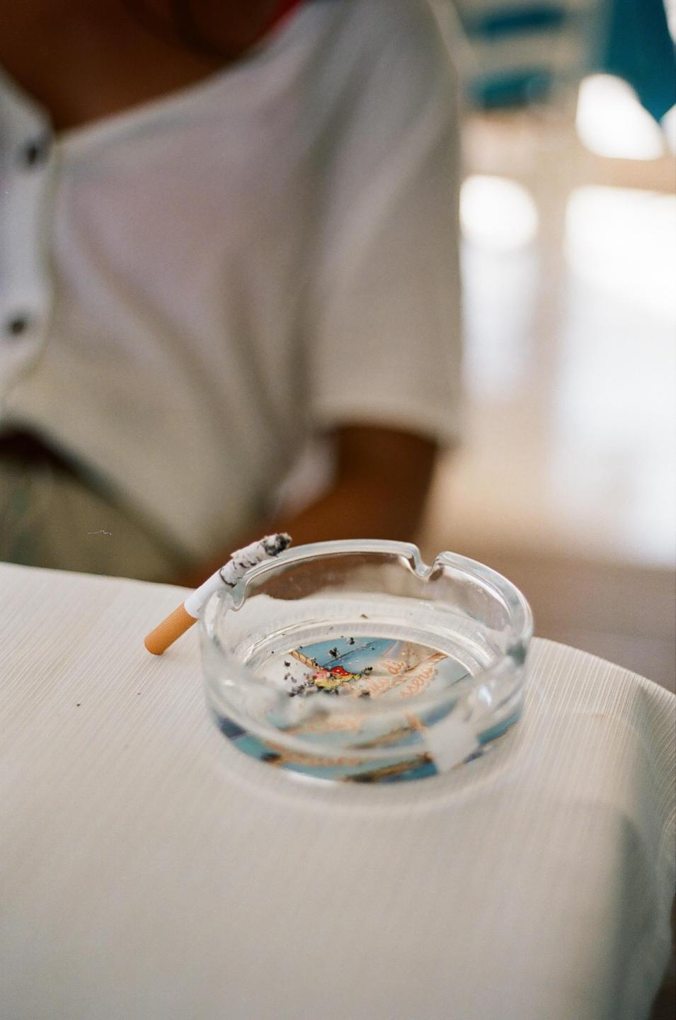A lit cigarette rests on a glass ashtray on a white table, with a person in a white shirt visible in the background.