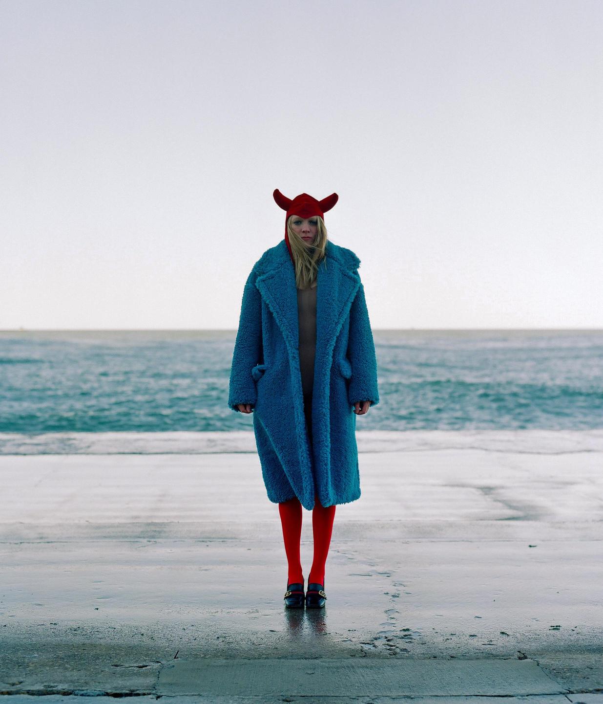 A woman in a blue coat, red tights, and a red horned hat stands on a wet concrete surface by the ocean.