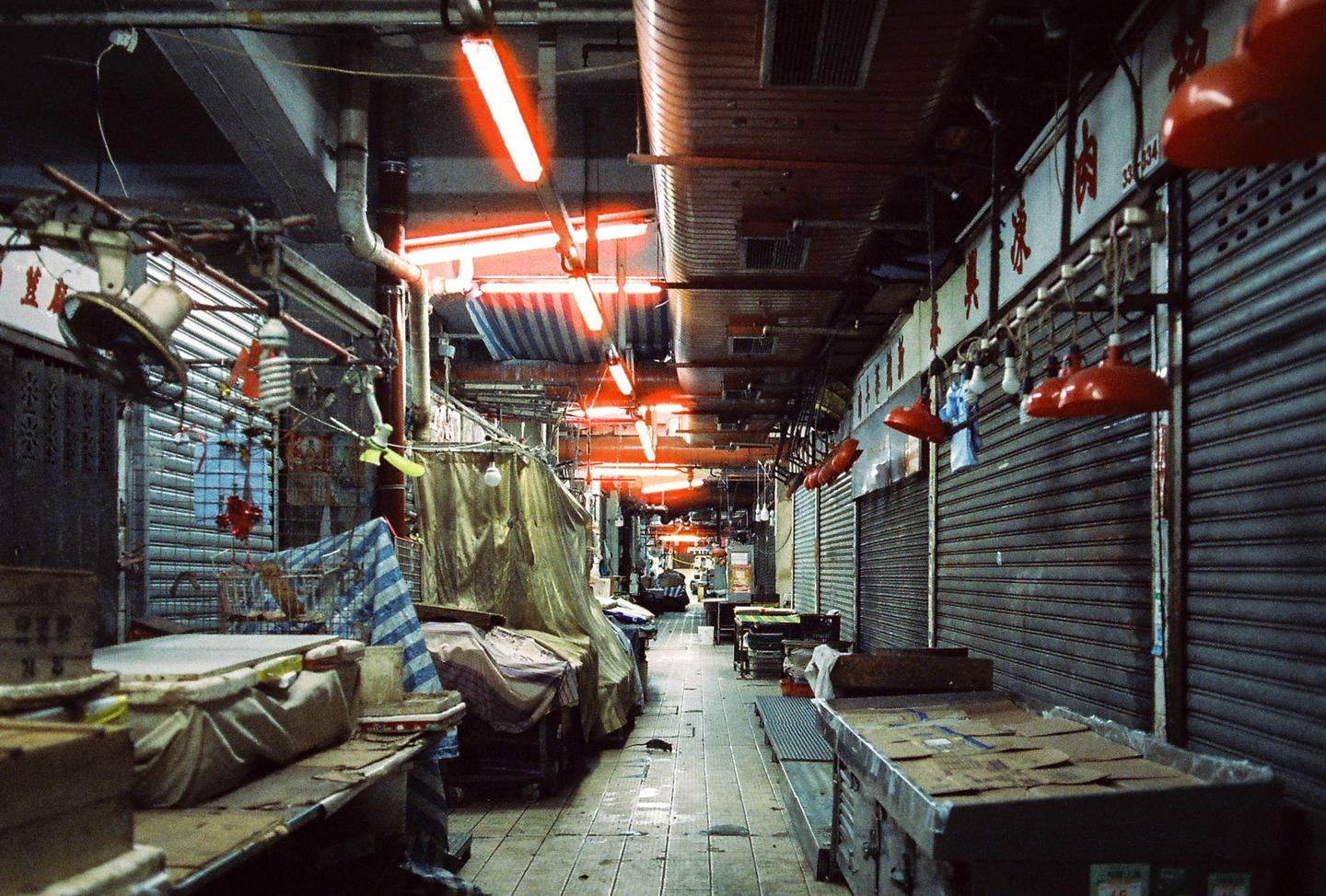 A narrow market alley is illuminated by red neon lights and red lamps, with metal stalls and closed metal shutters lining both sides.