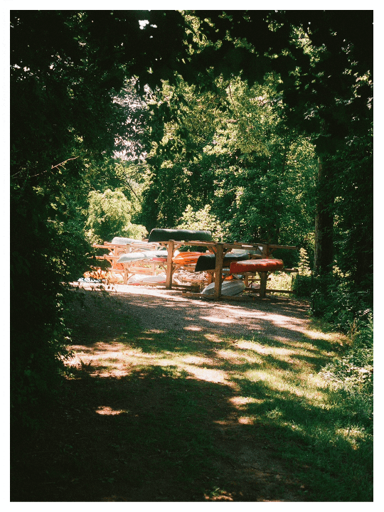 A rack of canoes and kayaks sits under a canopy of green trees, with dappled sunlight on the ground.