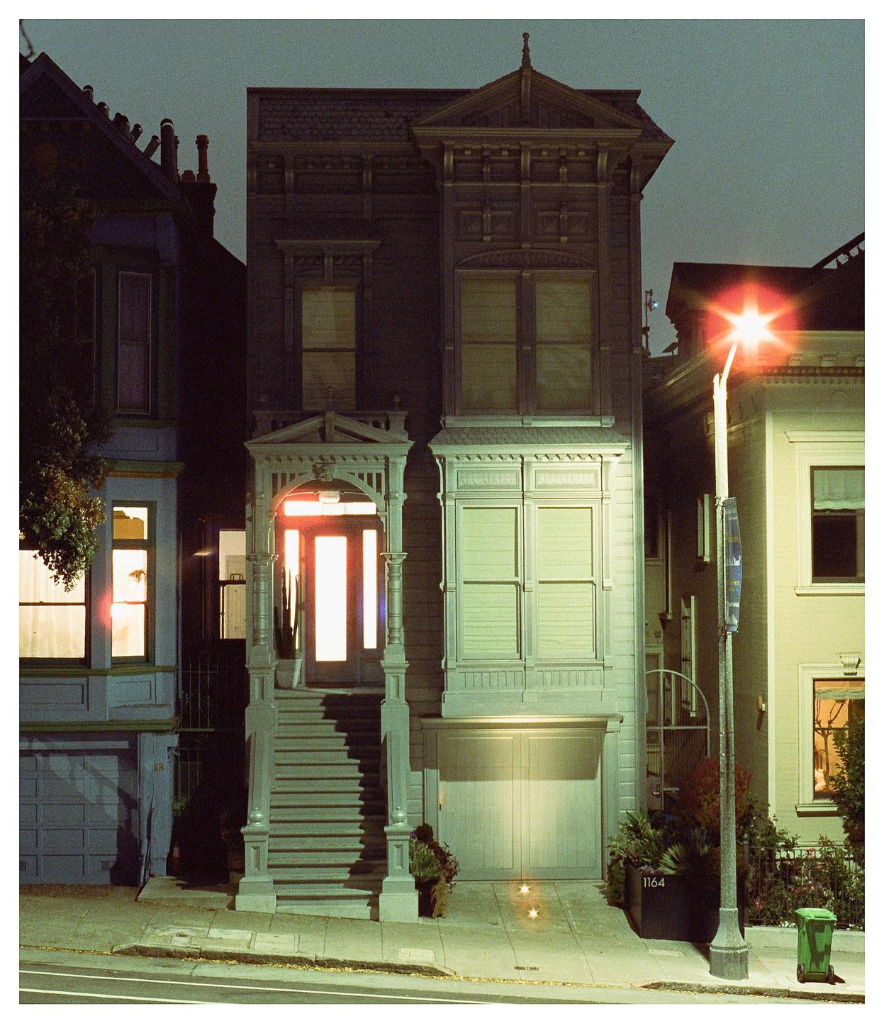 A Victorian house with a brightly lit doorway stands on a street at night, with a streetlamp glowing on the right.