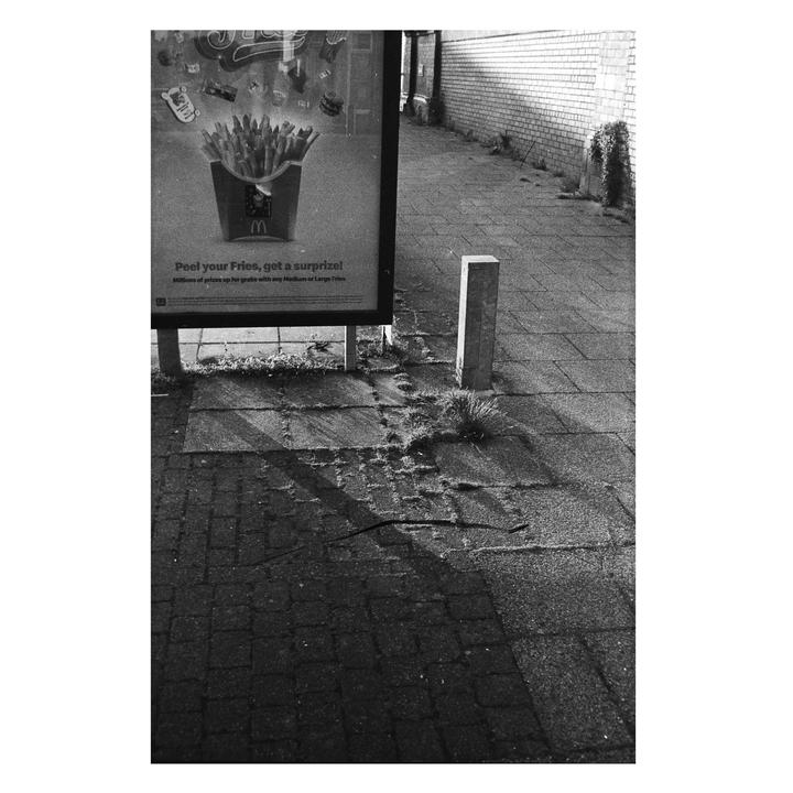 A black and white street photograph shows a billboard advertising french fries next to a brick wall and a sidewalk.