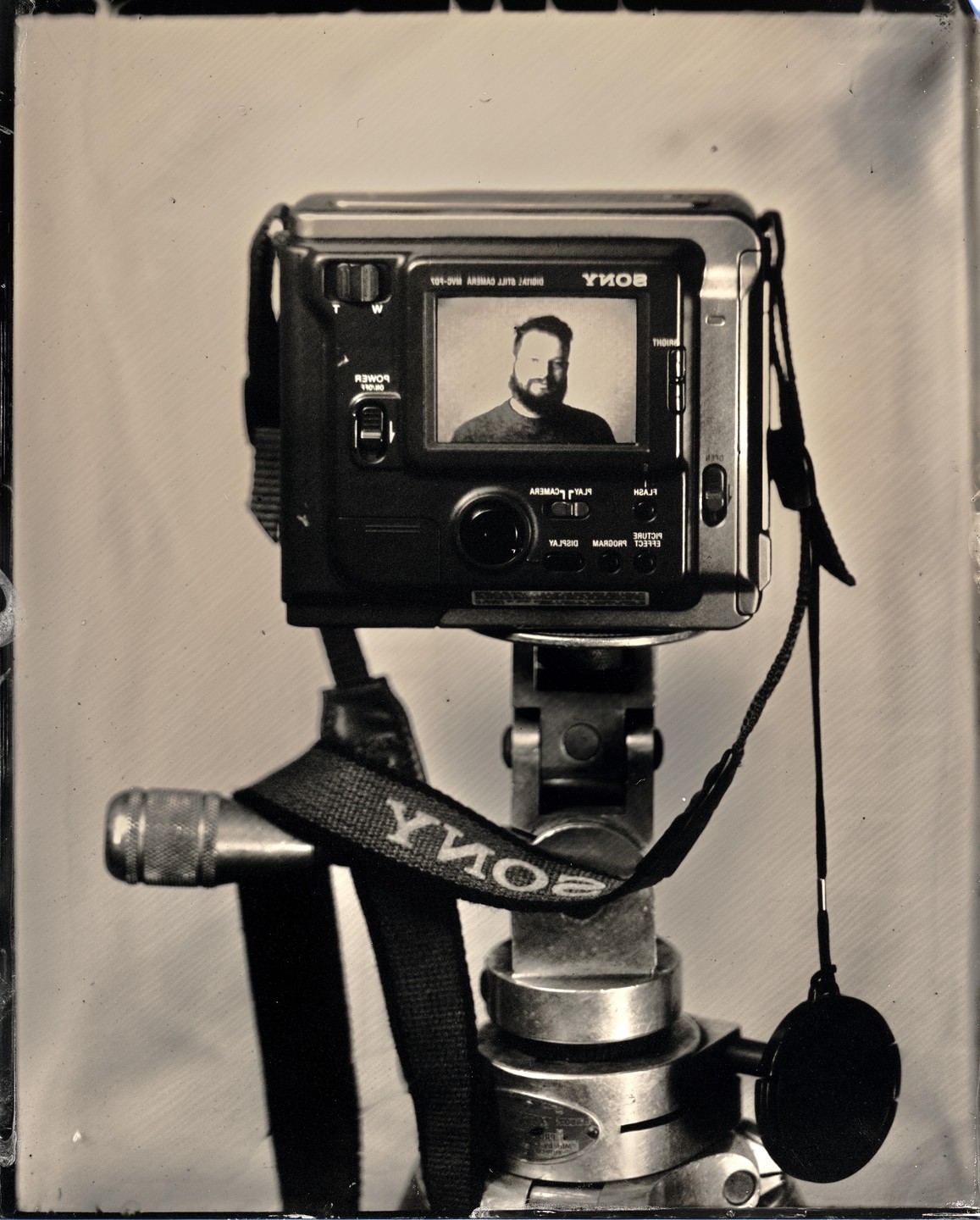 A digital camera on a tripod displays a self-portrait of a man with a beard on its screen.