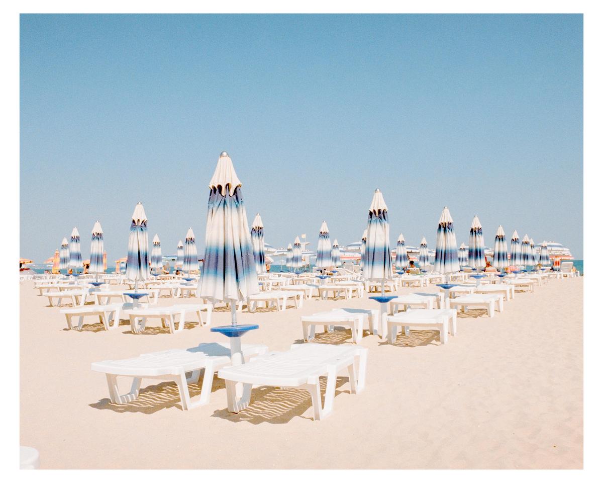 Many white and blue striped beach umbrellas and white lounge chairs are arranged in rows on a sandy beach under a clear sky.