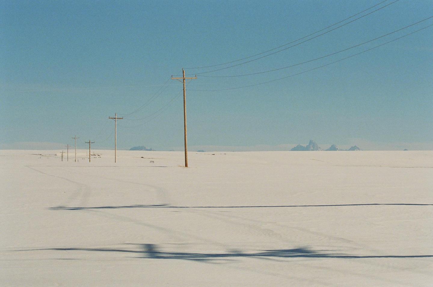 A vast, snow-covered plain stretches towards distant mountains under a clear sky, with a line of utility poles and their shadows extending into the distance.