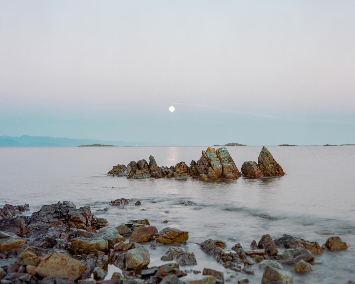 The moon is visible above a body of water with several rocky formations in the foreground and distant land in the background.