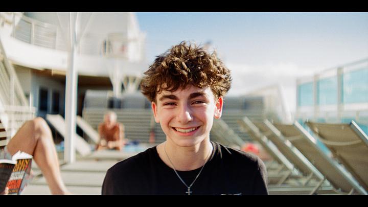 A young man with curly hair smiles at the camera while sitting outdoors on what appears to be a cruise ship deck.