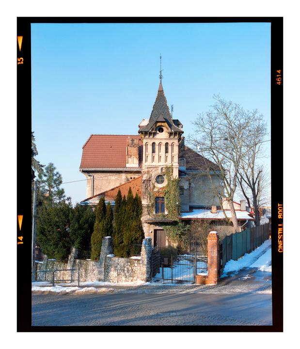 A large house with a prominent turret and red tile roof stands behind a stone fence and gate, with snow on the ground and bare trees.