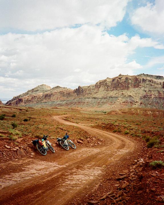Two bicycles are parked on the side of a winding dirt road in a desert landscape with mountains in the background.