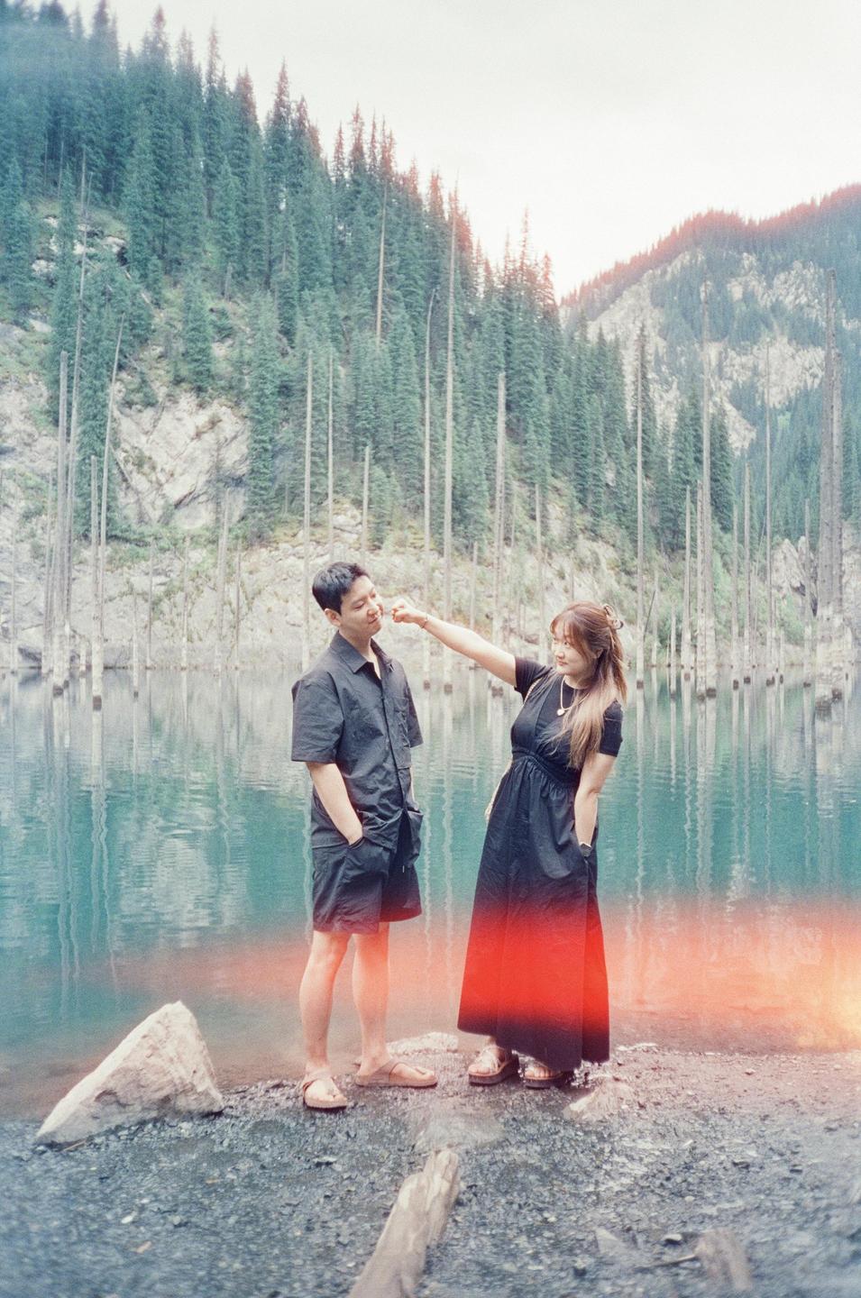 A couple stands by a lake with submerged trees and a forest on the mountain.