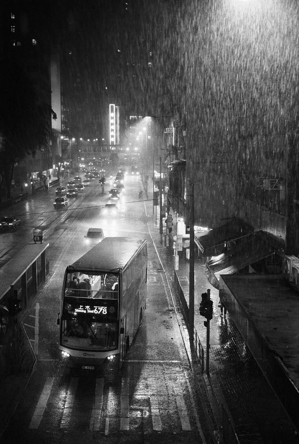A double-decker bus drives down a city street at night during a rainstorm.