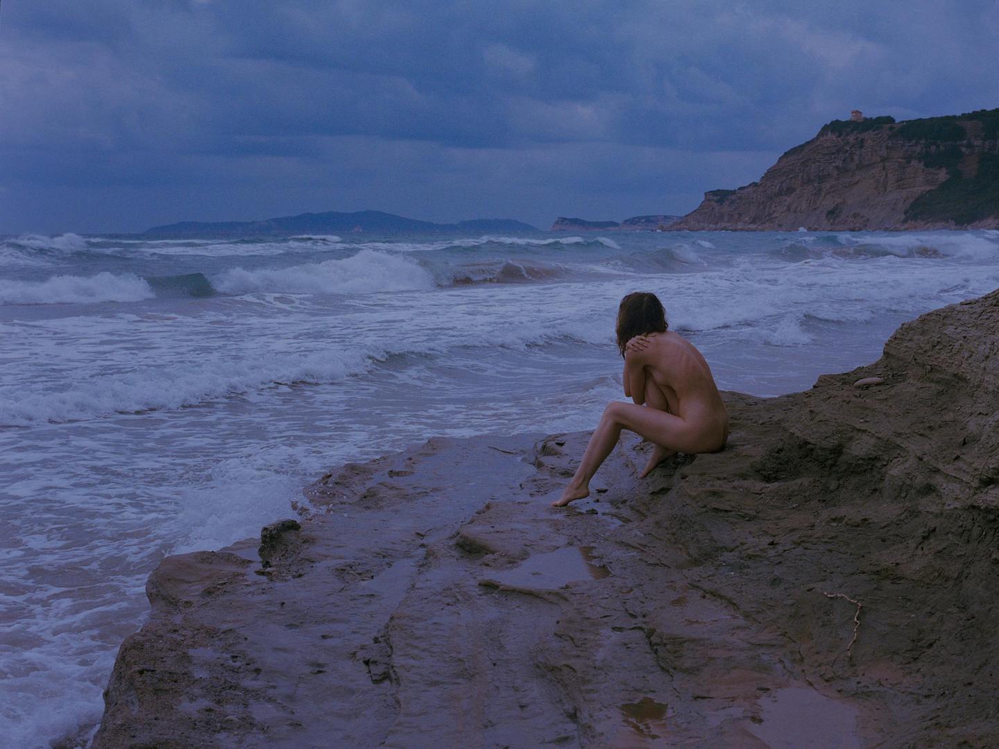 A nude woman sits on a rock by the ocean with waves crashing against the shore under a cloudy sky.