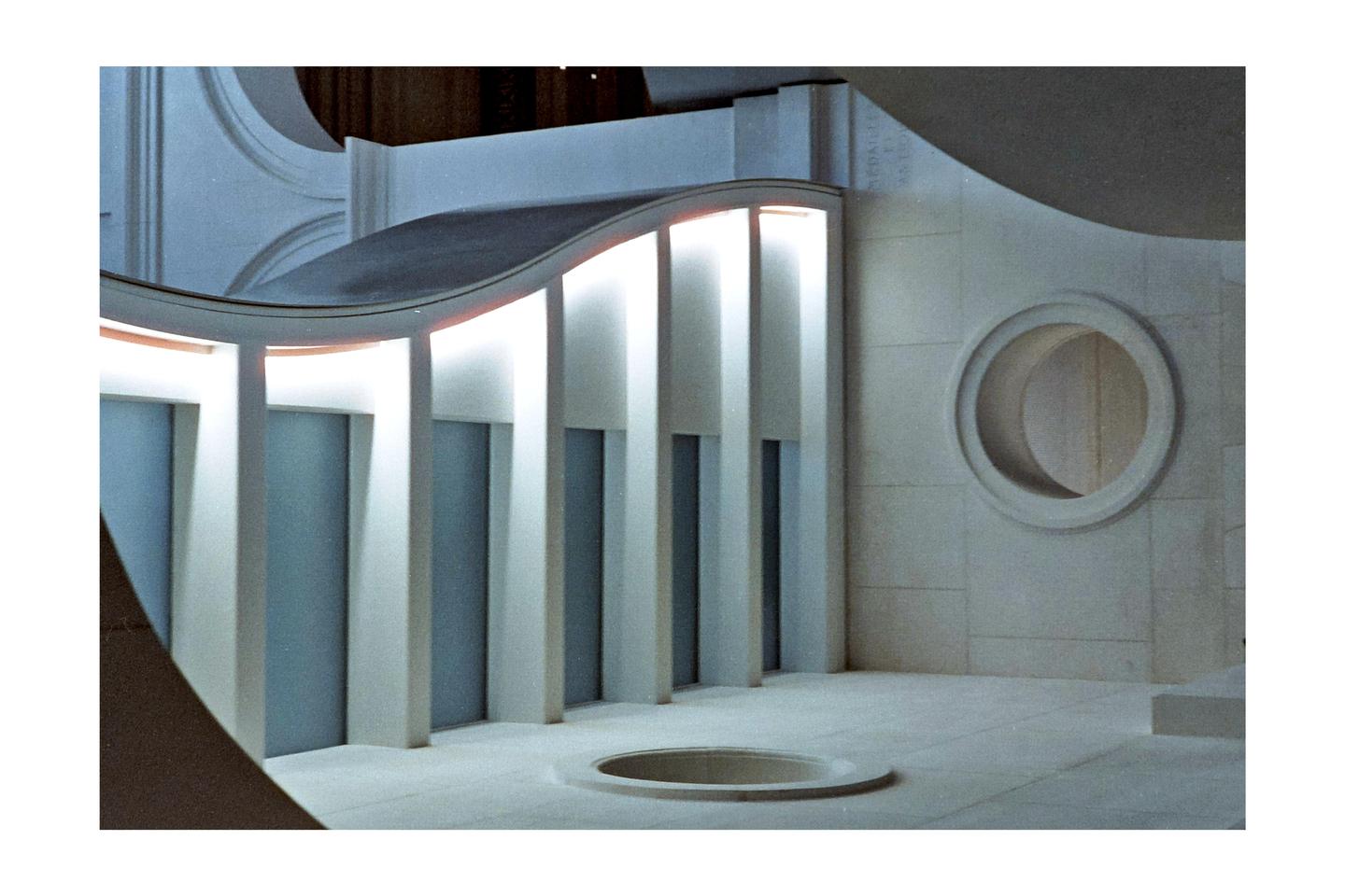 A model of a building with a curved roof, columns, and a circular window is illuminated by internal lights.