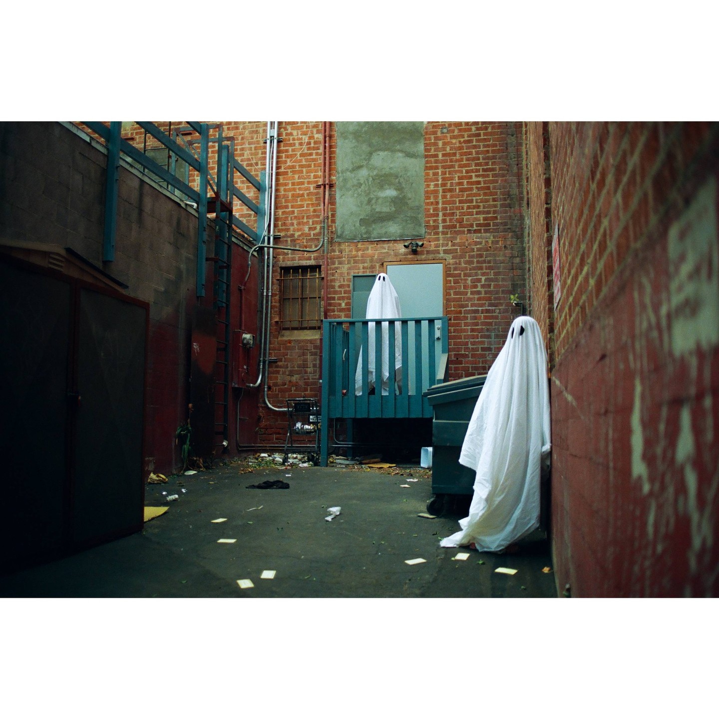 Two people dressed as ghosts stand in a brick alleyway next to a dumpster.