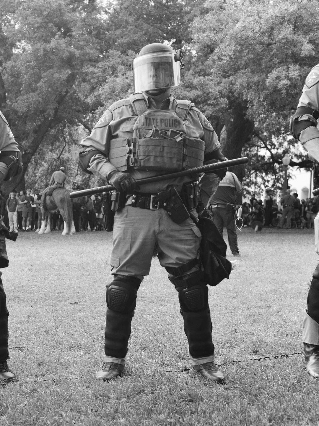 A police officer in riot gear stands on a grassy field holding a baton, with other people and trees in the background.