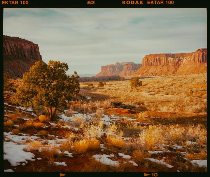 A desert landscape with red rock formations, sparse vegetation, and patches of snow is visible under a light sky.