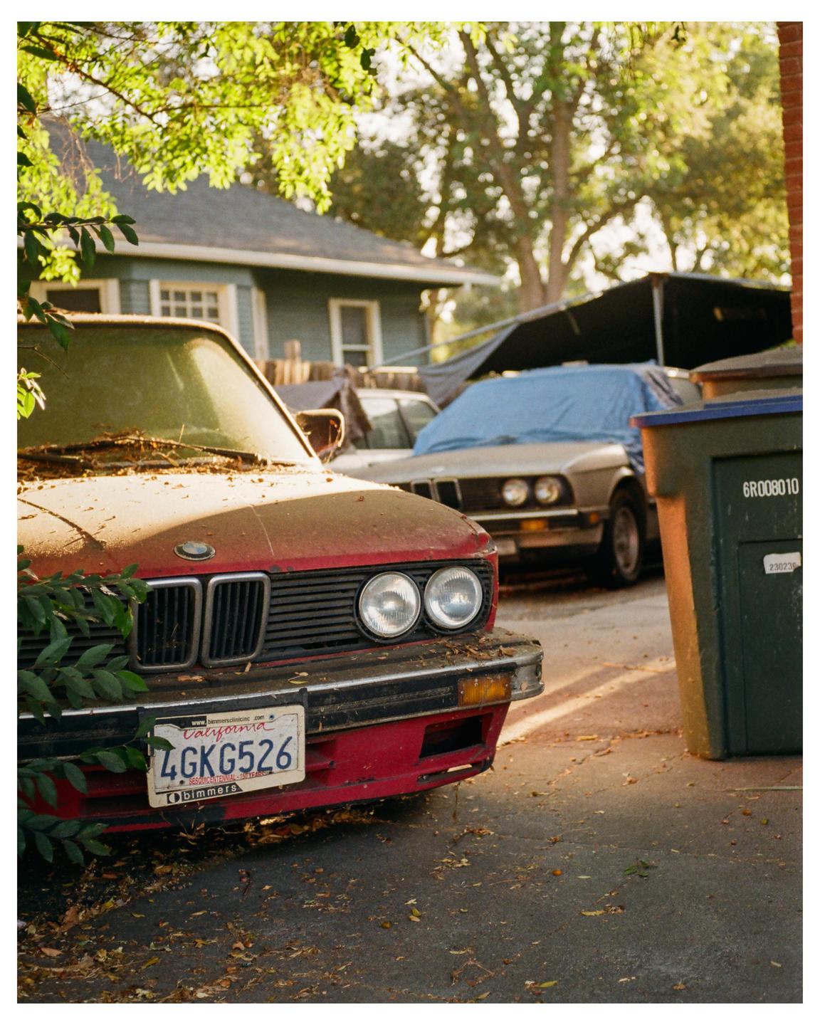 A dusty red car with a California license plate is parked in a driveway next to a green trash can and other cars.
