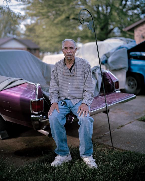 A man sits on the tailgate of a purple vehicle in a yard.