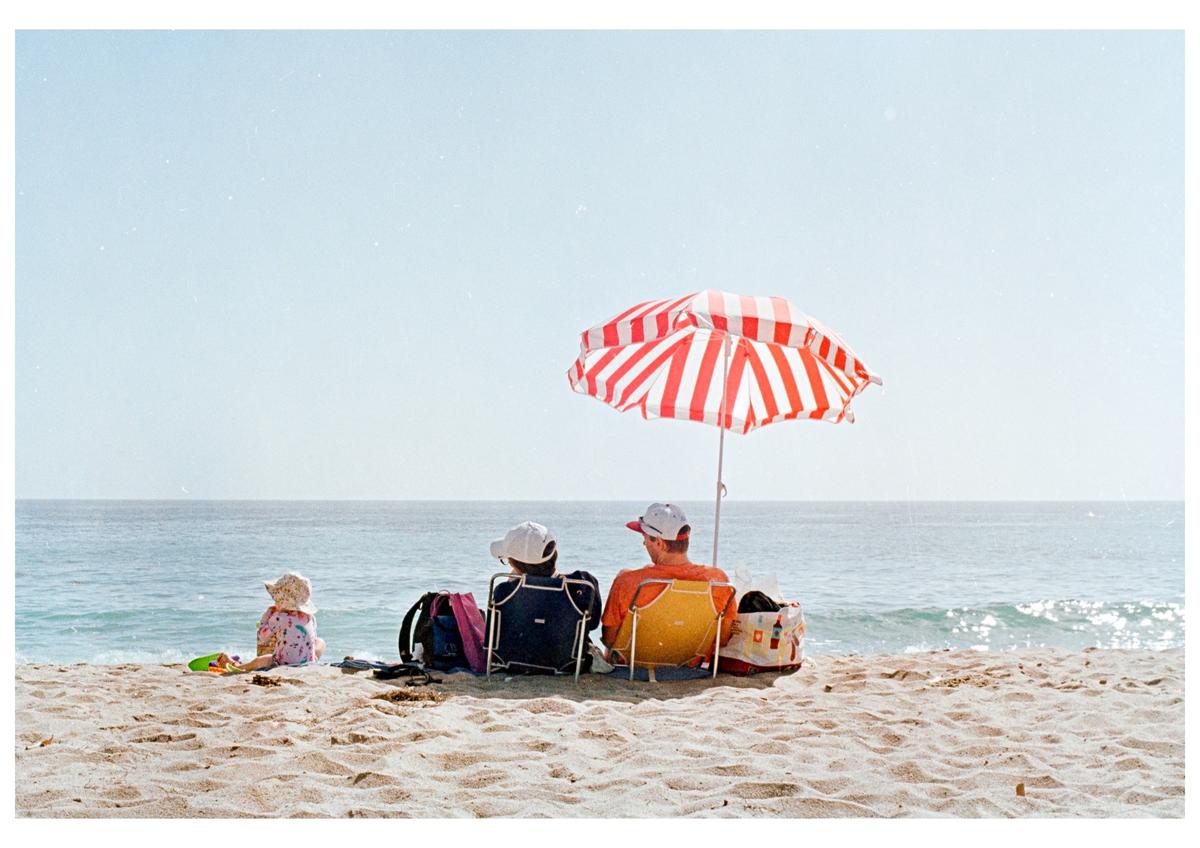 A family sits on a sandy beach under a red and white striped umbrella, looking out at the ocean.