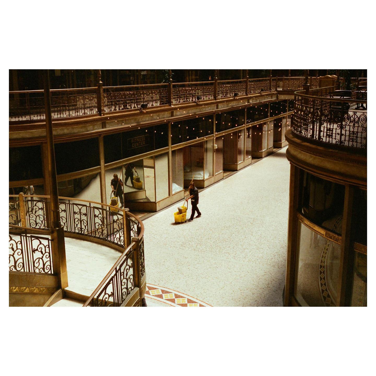 A man mops the floor of a multi-story indoor arcade with ornate railings and storefronts.