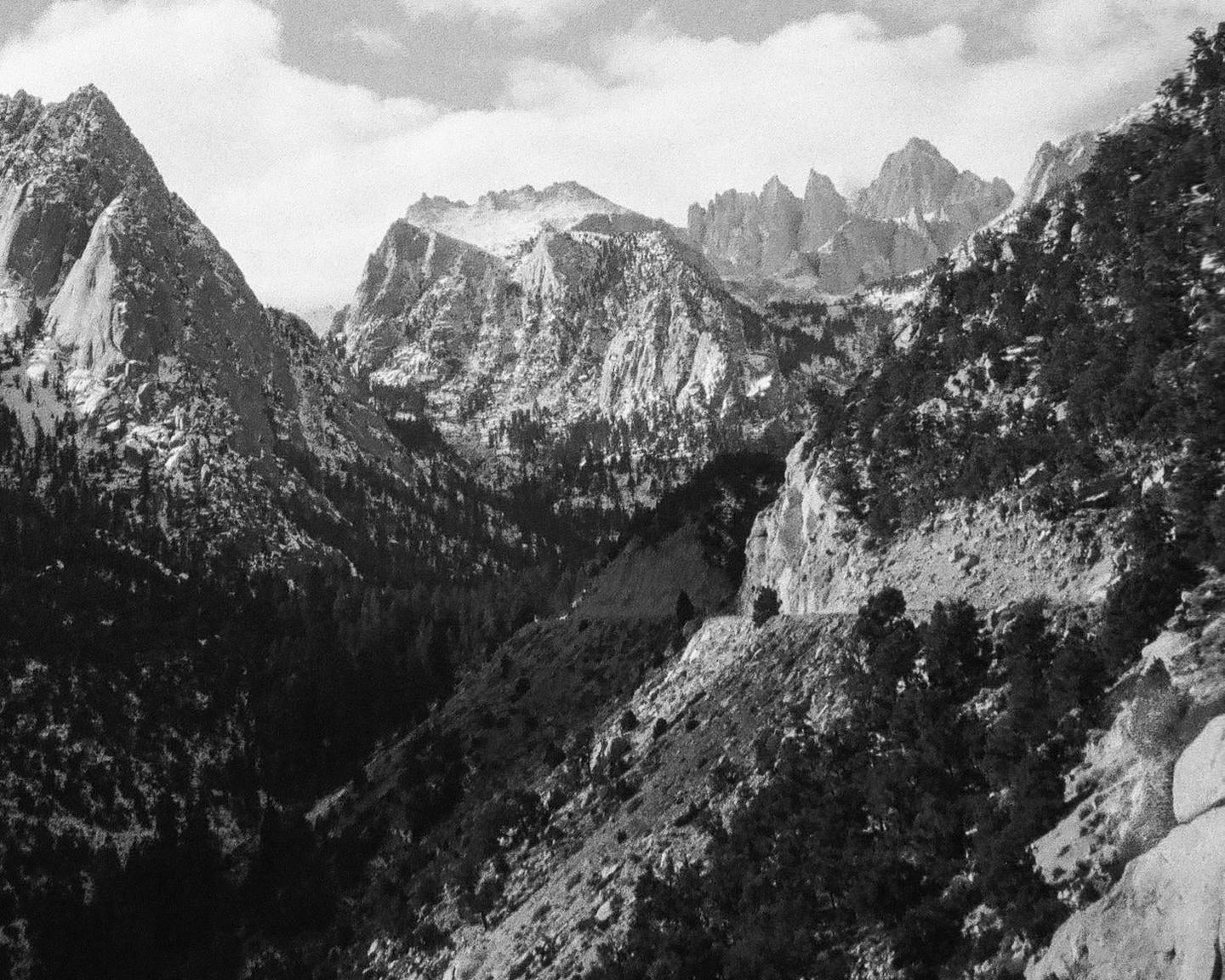 A black and white landscape shows a valley between two mountains with trees and clouds in the sky.