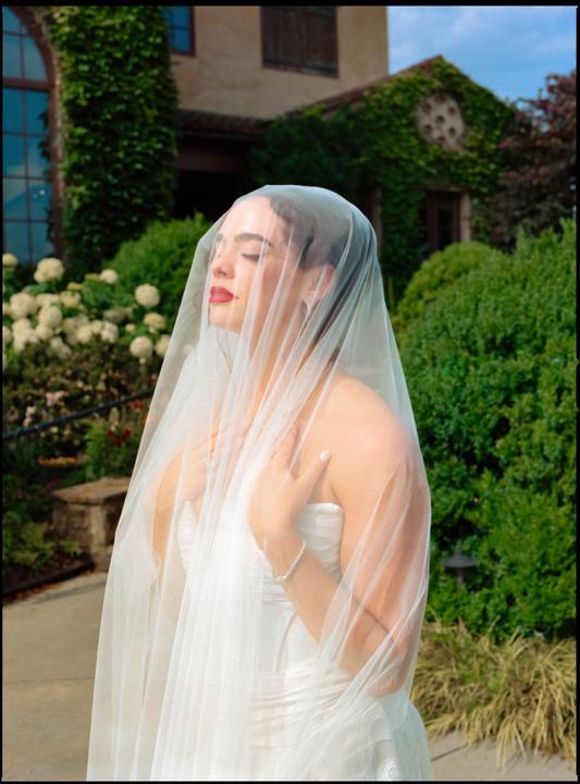 A woman in a wedding dress and veil stands with her eyes closed and hands on her chest in front of an ivy-covered building.