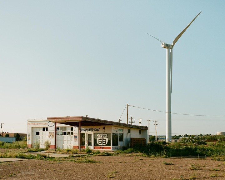 An abandoned gas station with a "Historic Route 66" sign stands next to a large wind turbine under a clear sky.