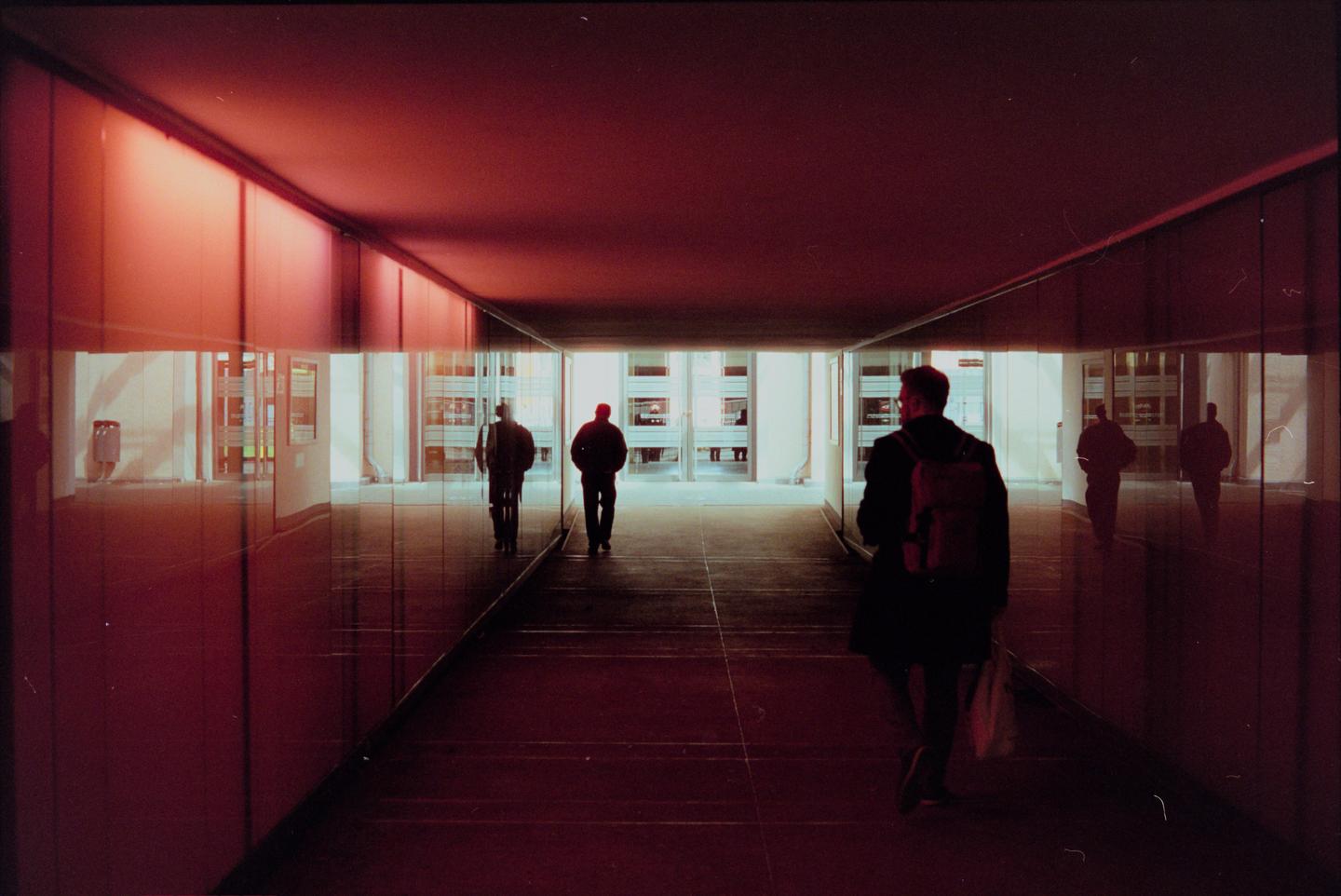 Three men walk through a red-lit tunnel with reflections on the walls.