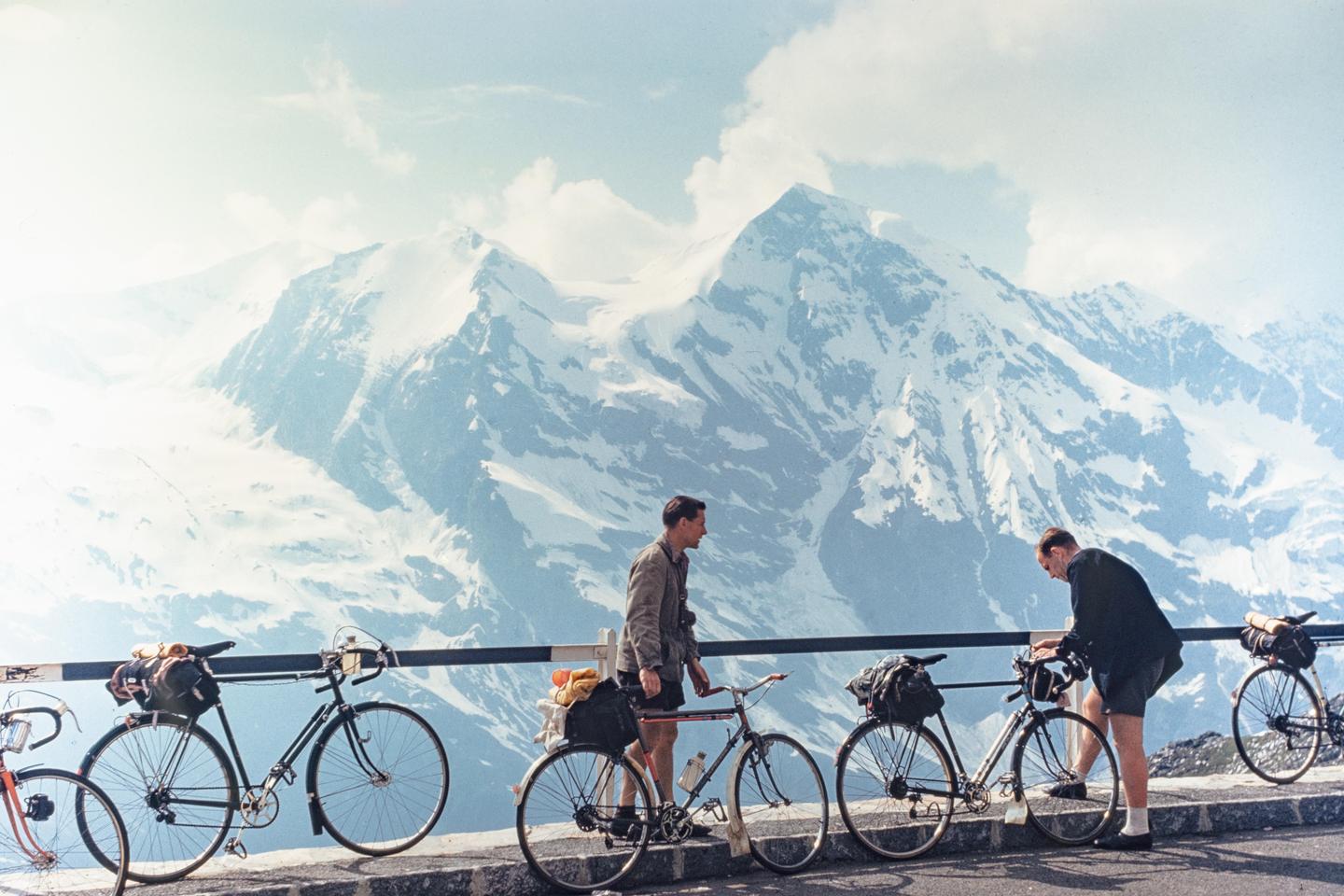 Two men stand with their touring bicycles on a mountain road overlooking snow-capped peaks.
