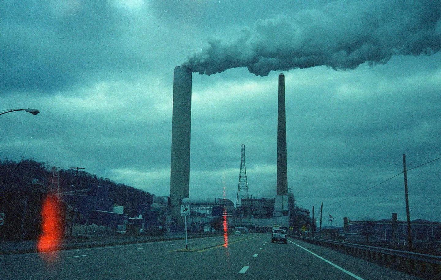 Smoke billows from two tall industrial smokestacks next to a highway under a cloudy sky.
