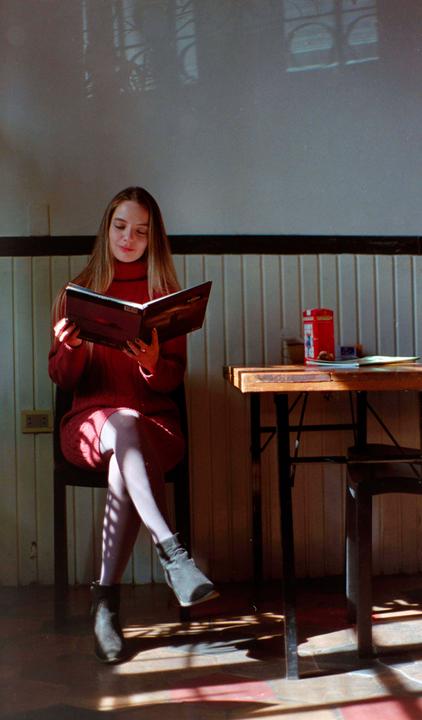 A woman in a red dress sits at a table and reads a book, with sunlight casting shadows on the floor.