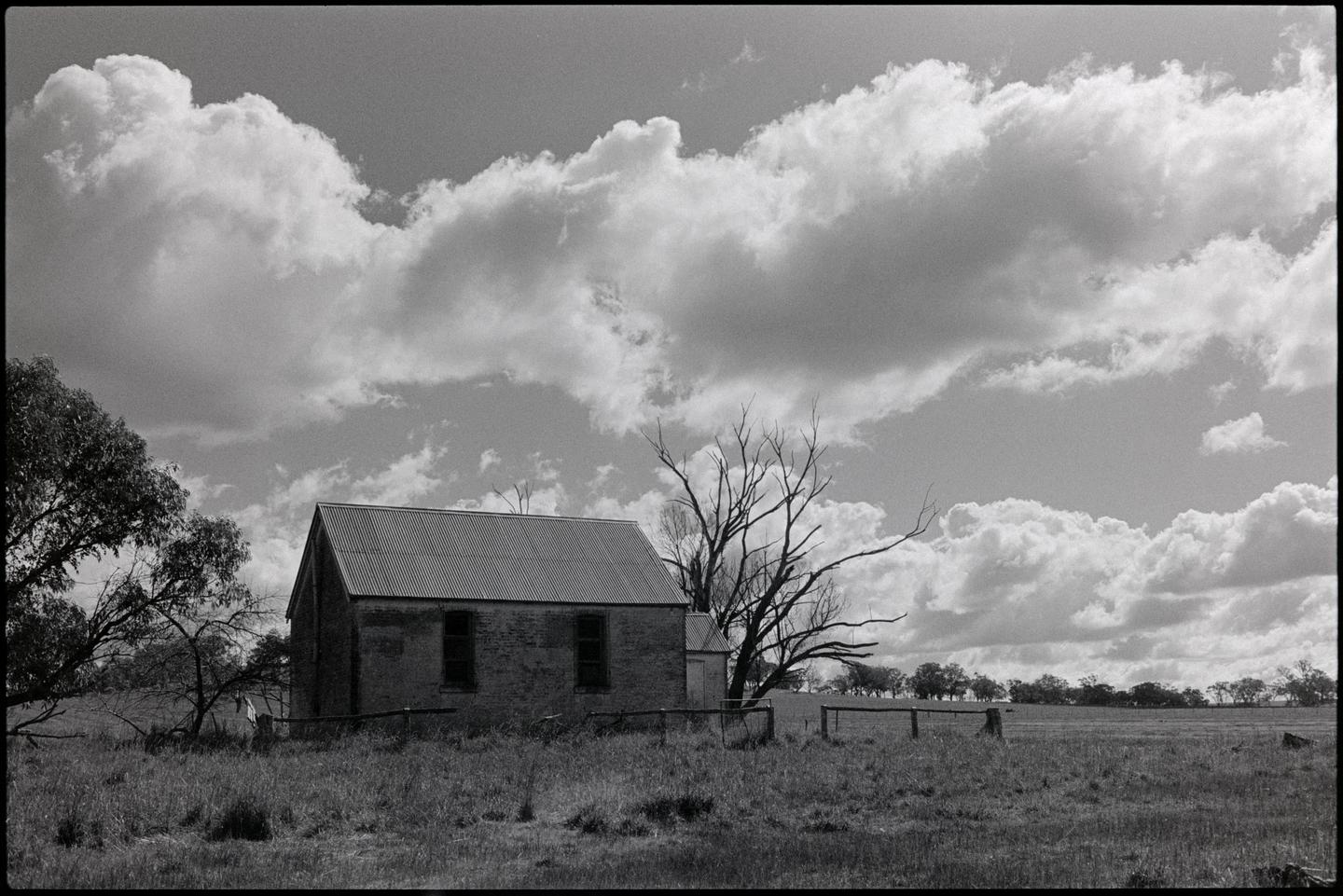 An old brick building with a corrugated iron roof stands in a grassy field under a cloudy sky.