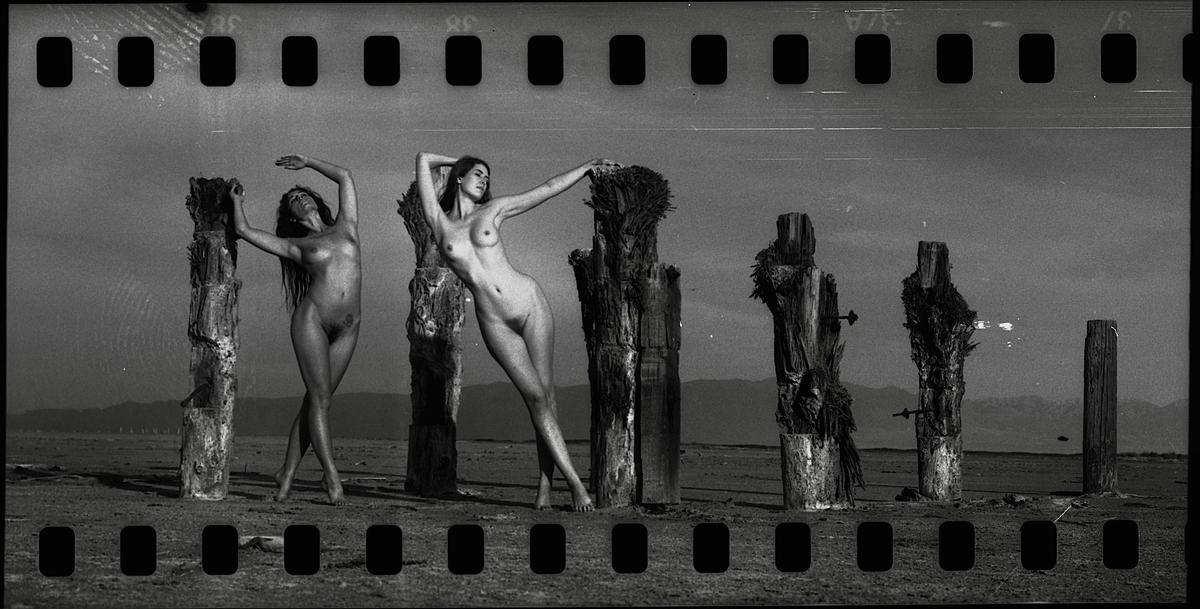 Two women pose nude next to weathered wooden posts in a barren landscape with mountains in the distance.