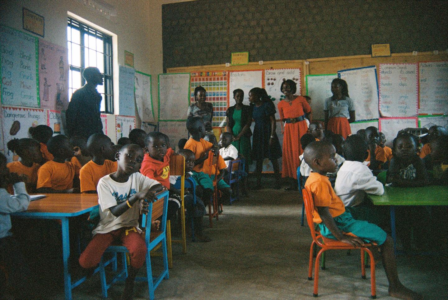 A group of children and adults are gathered in a classroom with educational posters on the walls.