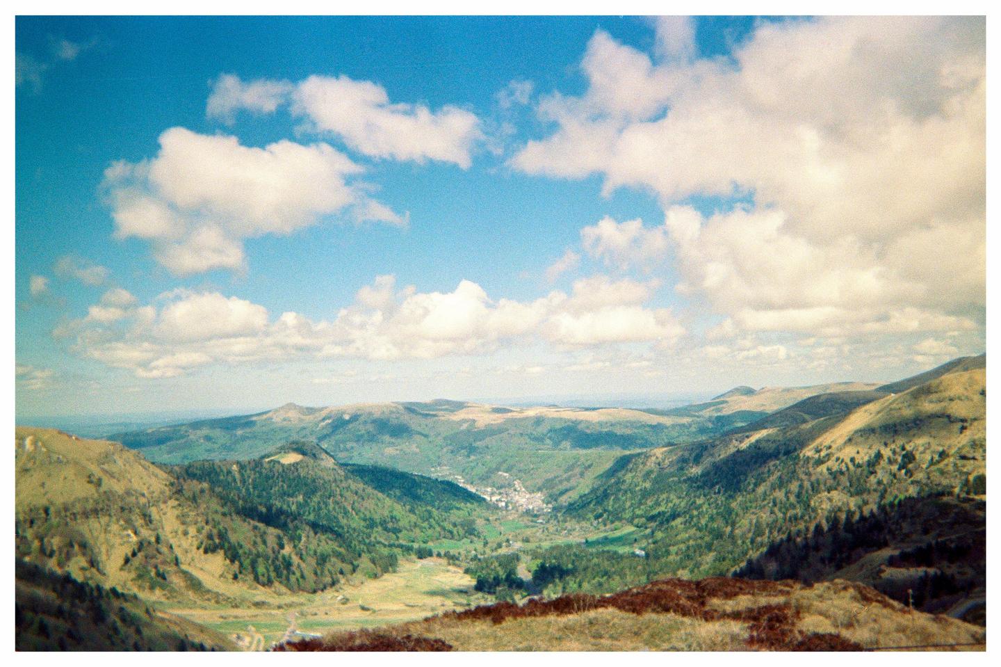 A small village is nestled in a valley surrounded by rolling green hills under a blue sky with white clouds.