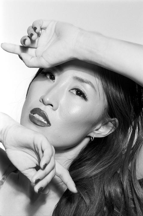 A woman with long hair and dark lipstick poses with her hands near her face.