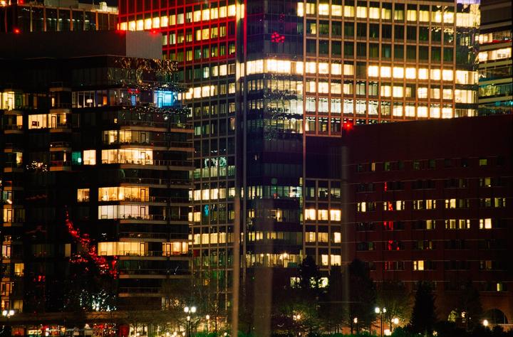 A cluster of illuminated buildings with many windows is visible at night, with trees and streetlights in the foreground.