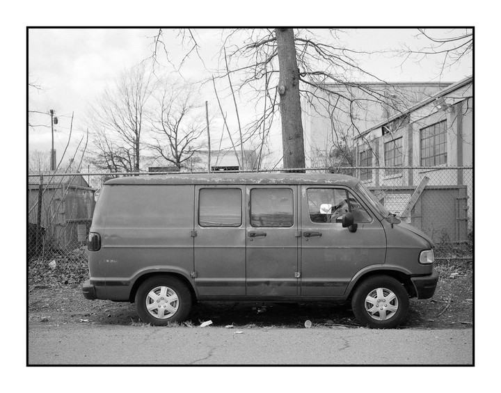 A gray van is parked on a street next to a chain-link fence and a building with many windows.