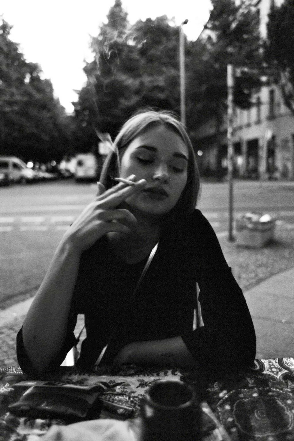A woman with closed eyes smokes a cigarette at a table on a street.