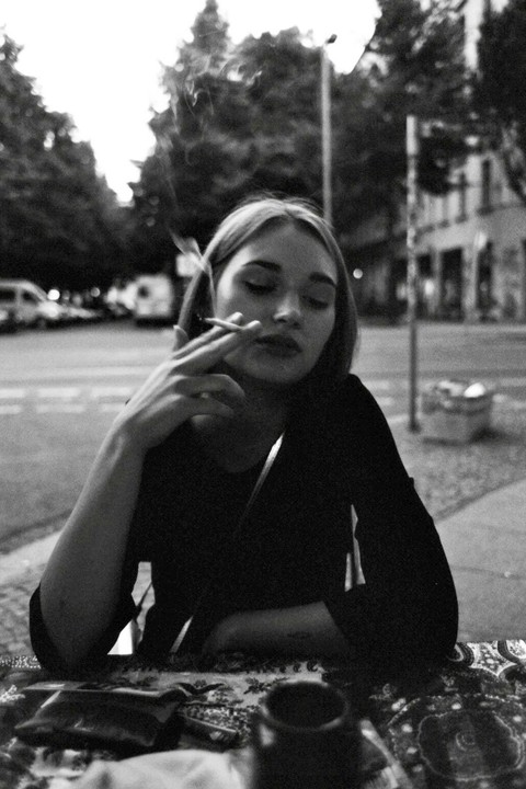 A woman with closed eyes smokes a cigarette at a table on a street.
