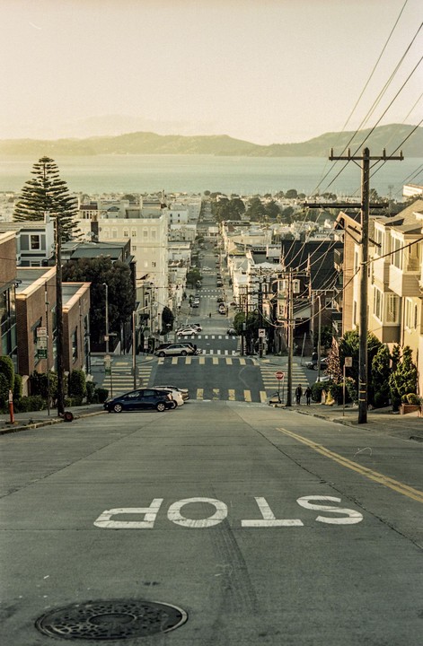 A steep street with a stop sign painted on the asphalt leads down to a body of water with hills in the distance.