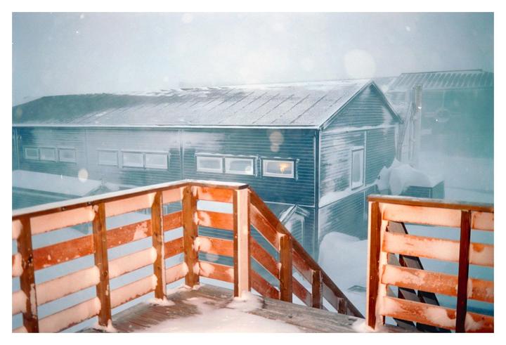 A snow-covered wooden deck and railing are in the foreground, with a building and snowdrifts in the background during a snowstorm.