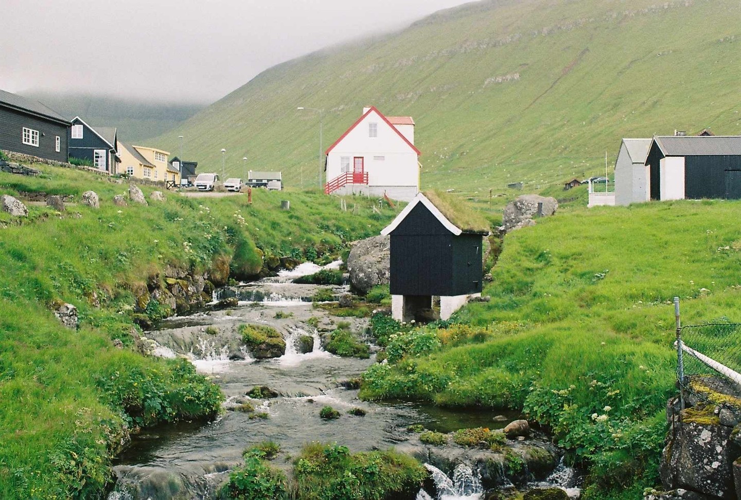 A small black house with a grass roof stands next to a cascading stream in a green valley with other houses and a road.