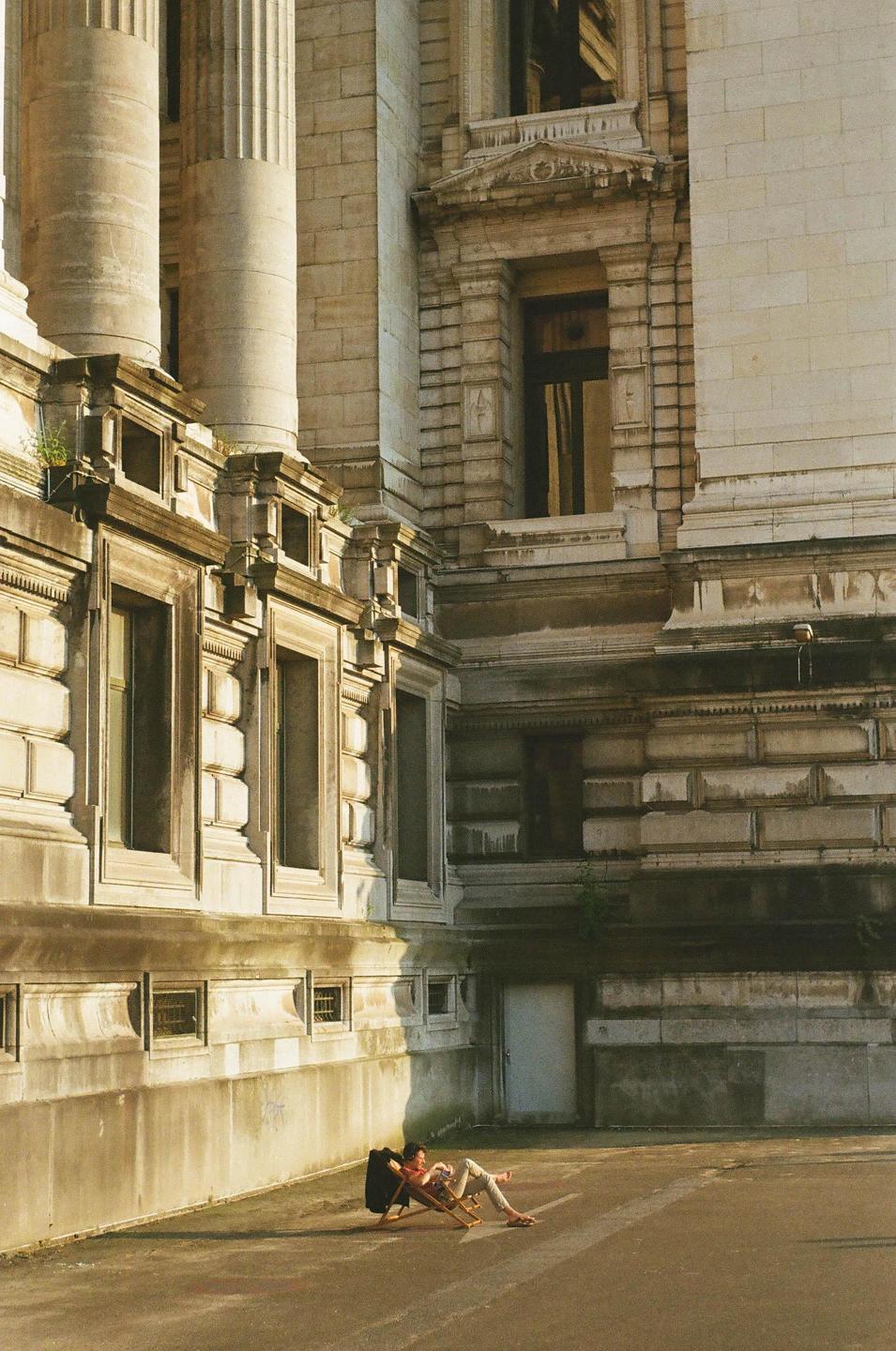 A woman relaxes in a folding chair on a paved area next to a large stone building with columns and ornate windows.