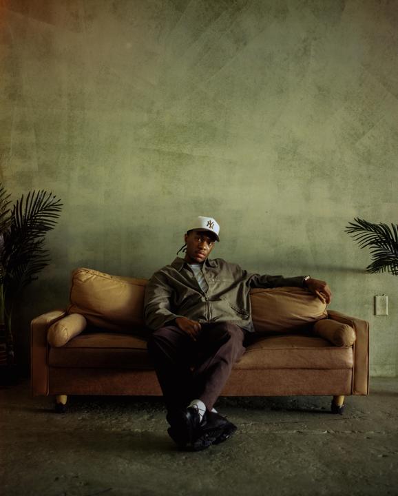 A man wearing a baseball cap and jacket sits on a brown sofa with plants on either side.